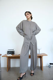 Woman in a gray outfit standing in a minimalistic room with a wooden table and white wall.